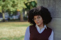 a young woman wearing glasses and a vest leaning against a wall